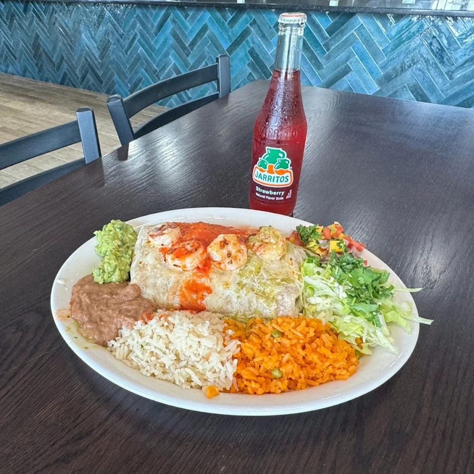 A shrimp burrito with rice, beans, guacamole, and salad, served with a Jarritos soda.