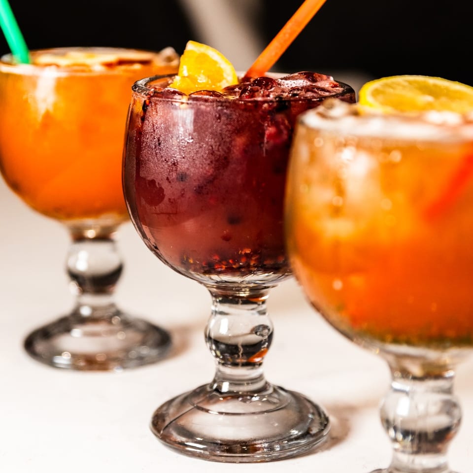 Three vibrant Mexican cocktails in stemmed glasses, two orange and one dark red, garnished with citrus.