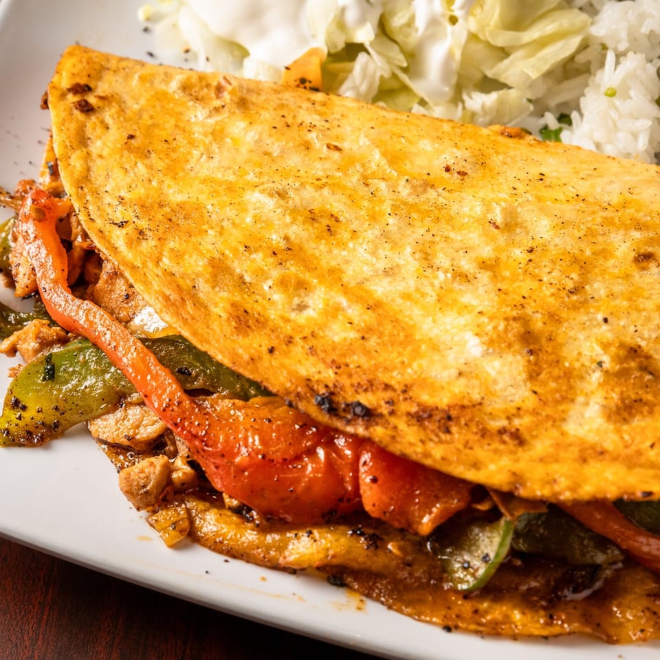 Close-up of a golden quesadilla filled with chicken, red and green bell peppers, served with rice and salad.